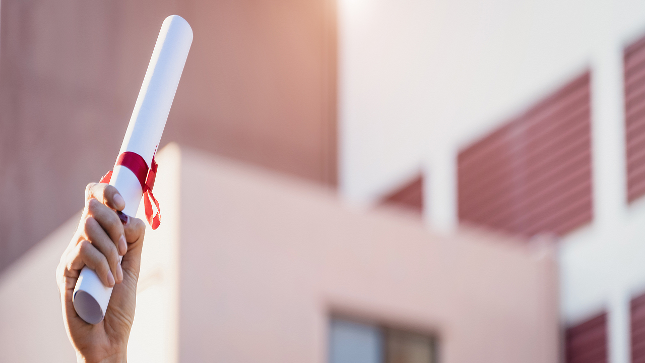 Hand holding a rolled diploma in the air with a building in the background